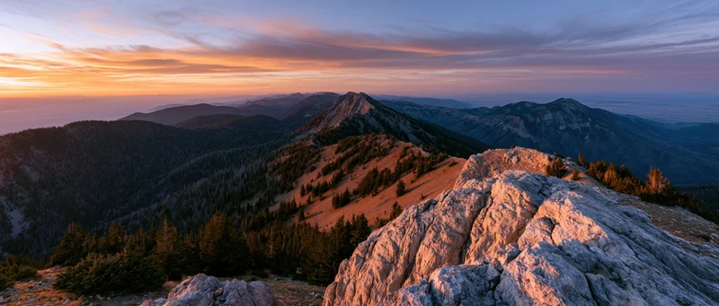 Alpine mountain panorama from rocky summit at sunset with forested valleys and colorful sky