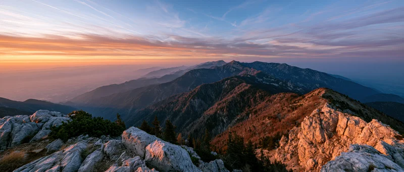 Alpine mountain range viewed from rocky summit at sunset with forested valleys and layered peaks under colorful sky