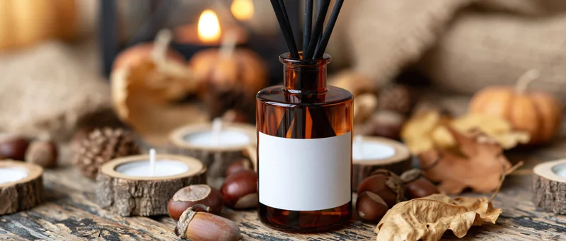Amber glass reed diffuser with blank label on rustic wooden table surrounded by autumn decor and lit candles