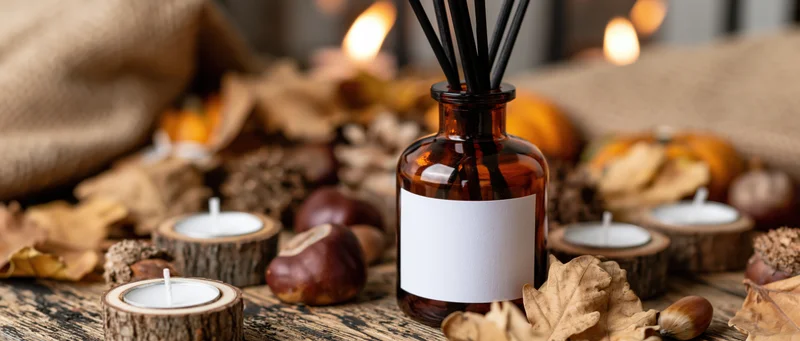 Amber glass reed diffuser with blank label surrounded by autumn leaves and candles on wooden table