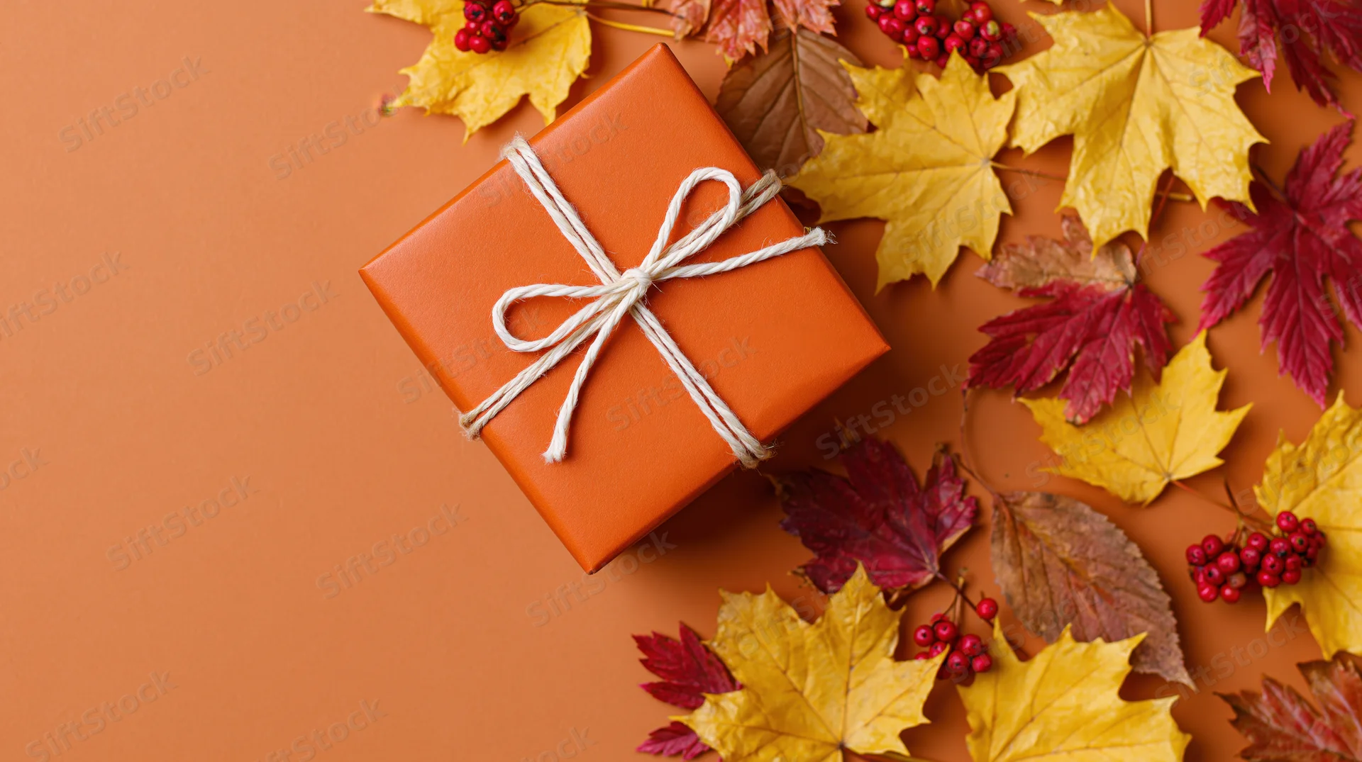 Orange gift box tied with white twine bow surrounded by autumn maple leaves and red berries on orange surface