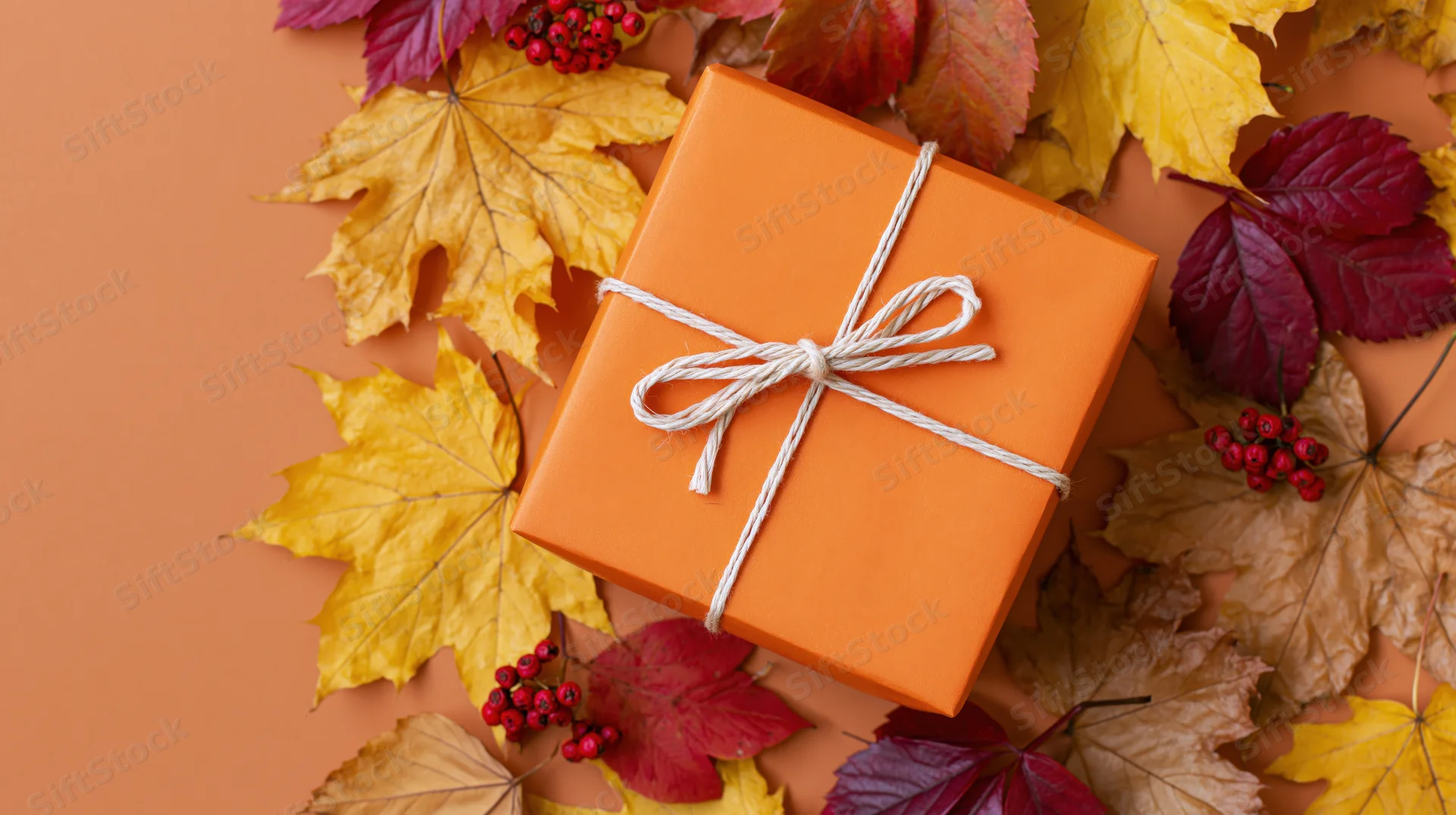 Orange gift box tied with white twine bow surrounded by autumn leaves and berries on warm-toned surface