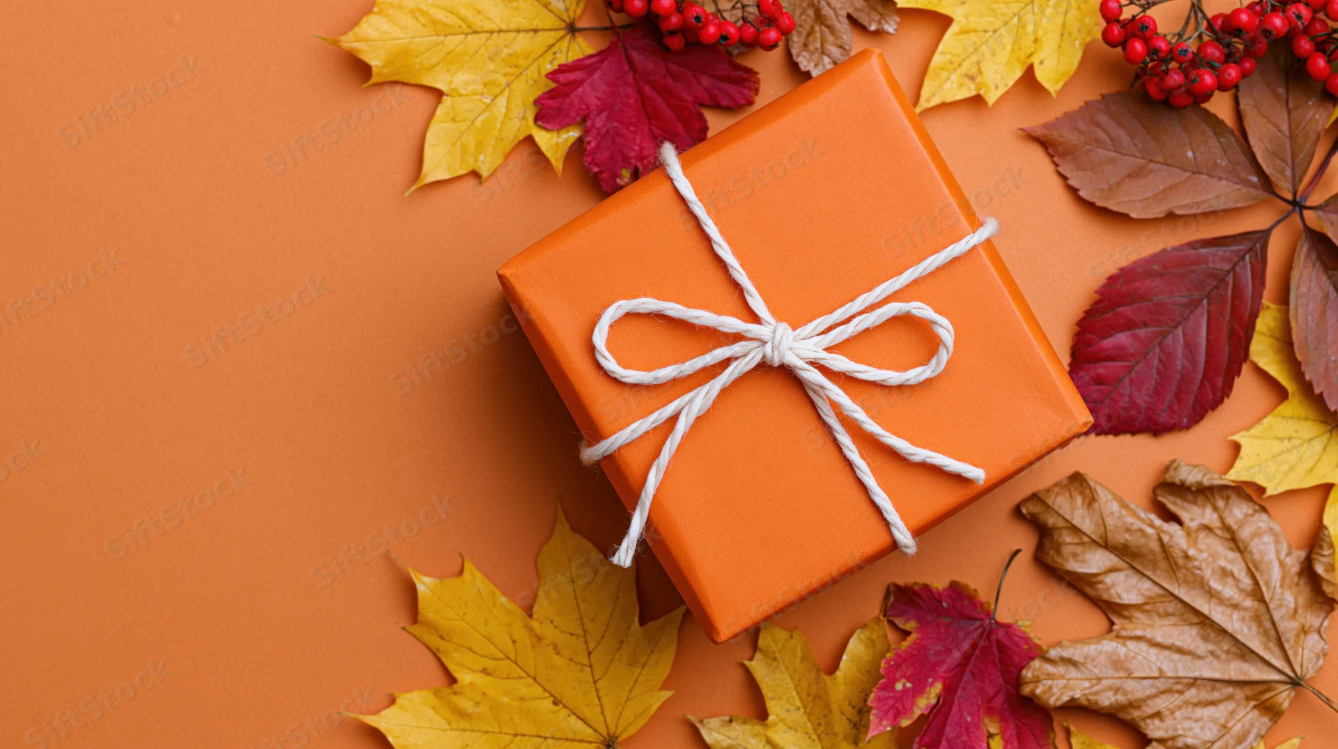 Orange gift box tied with white twine bow surrounded by autumn leaves on orange surface