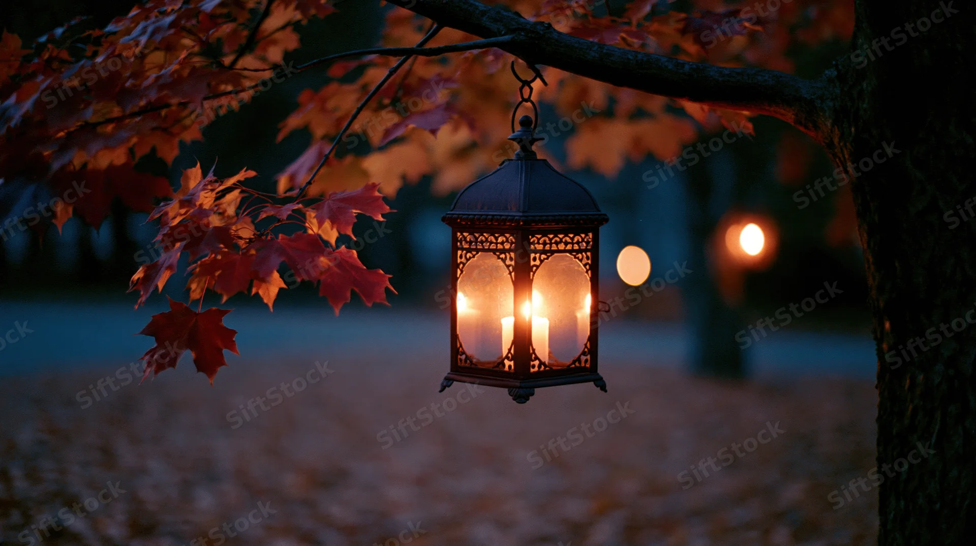 Ornate metal lantern glowing with warm candlelight hanging from a tree branch in an autumn garden at twilight