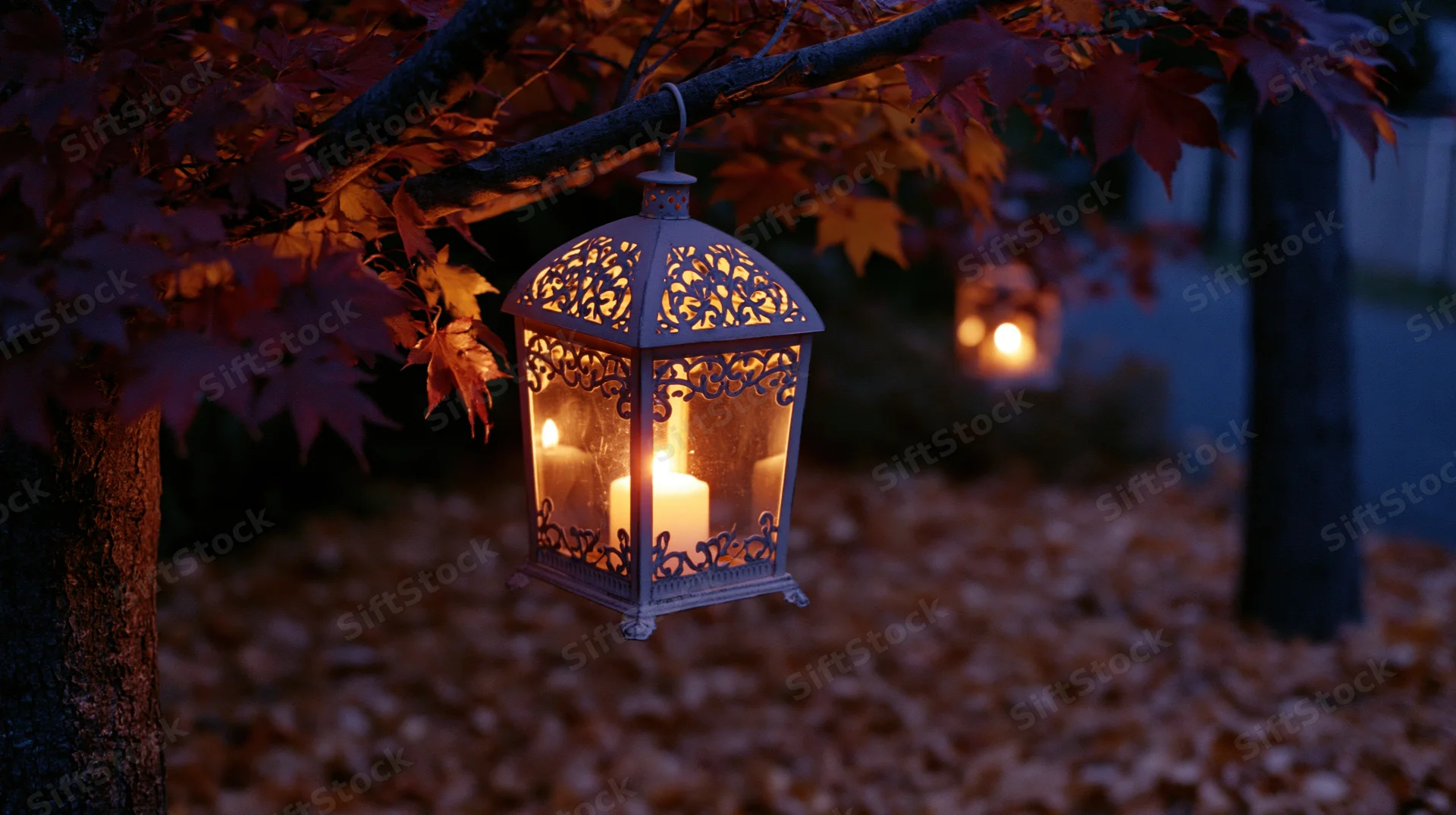 Ornate metal lantern glowing with candlelight hanging from tree branch in autumn garden at dusk