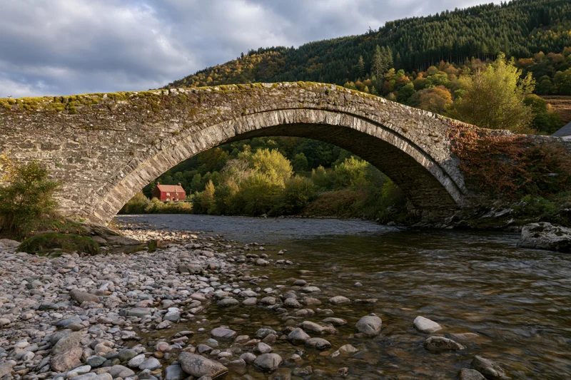 Ancient stone arch bridge spanning a shallow rocky river in a forested countryside landscape