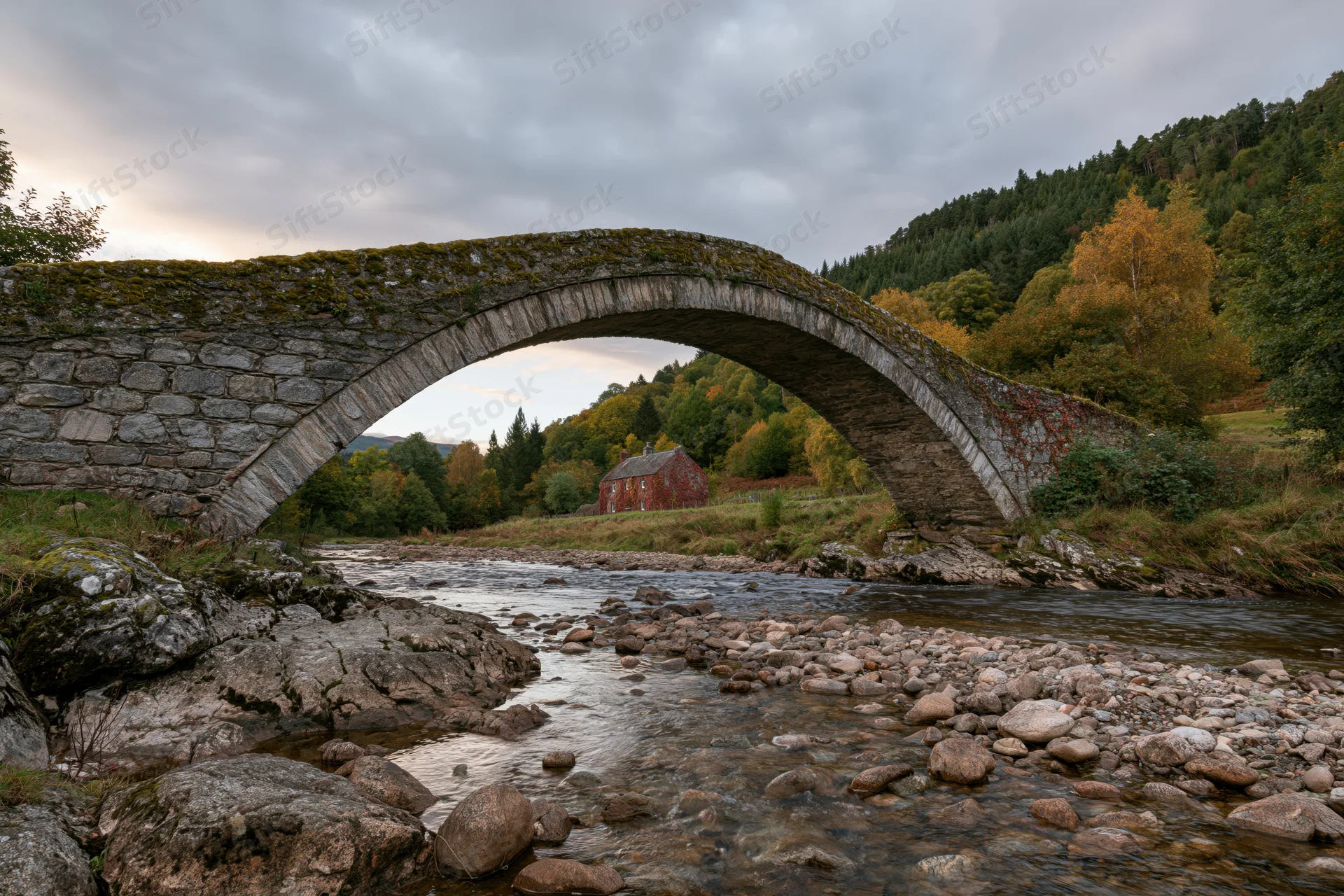 Ancient stone arch bridge spanning a shallow rocky river in a forested countryside landscape
