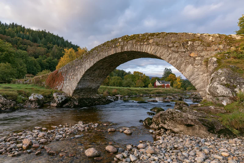 Ancient stone arch bridge spanning a shallow rocky river in a rural landscape with autumn trees and a distant house