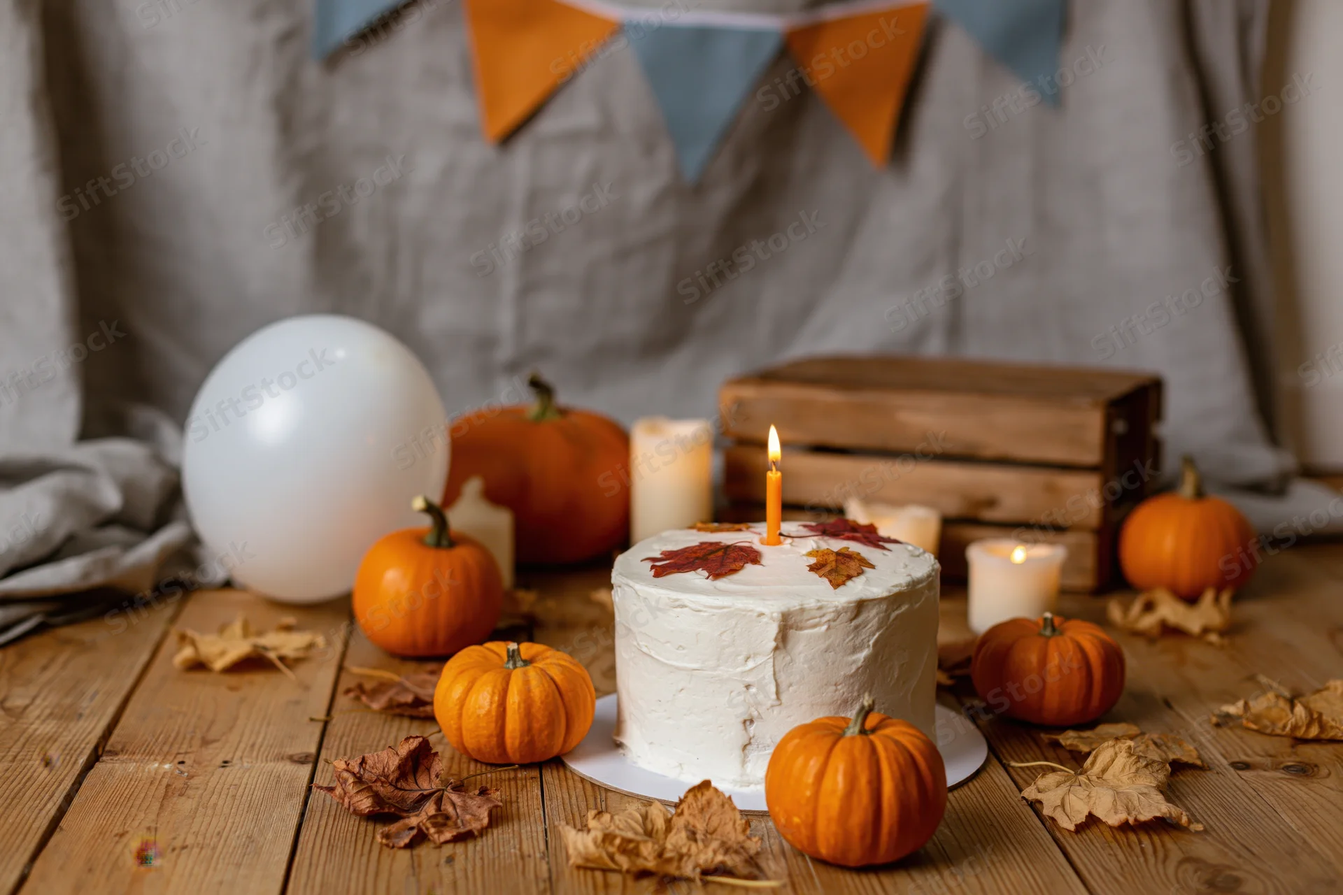 White frosted cake with lit candle surrounded by pumpkins and autumn leaves on wooden floor