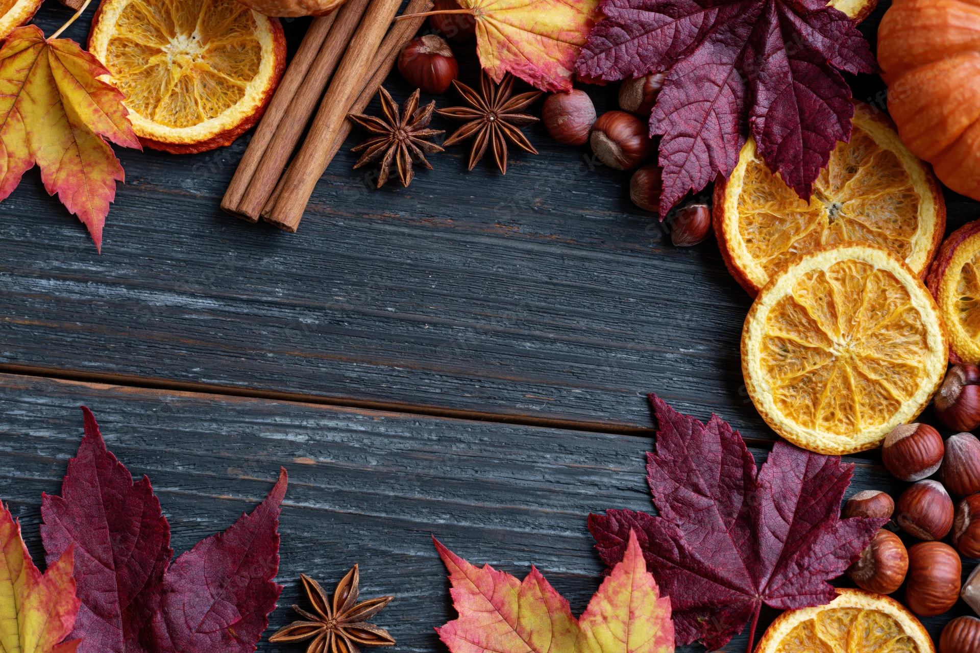Autumn flat lay with dried orange slices, cinnamon sticks, maple leaves, hazelnuts, and star anise arranged on dark wooden surface with central copyspace