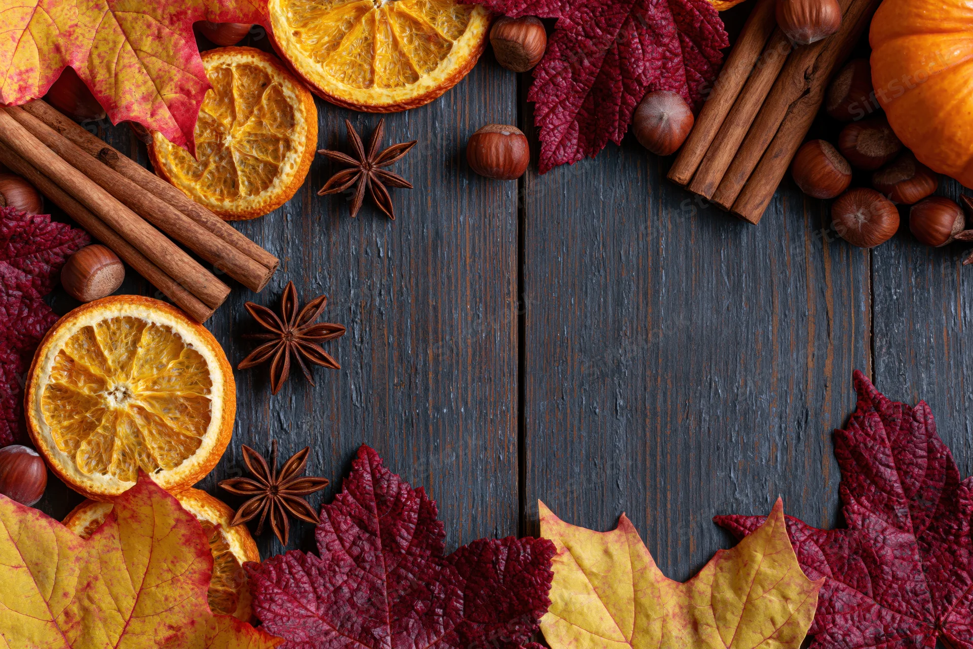 Autumn flat lay arrangement of dried orange slices, cinnamon sticks, star anise, hazelnuts, and colorful maple leaves on dark wooden table with central copyspace