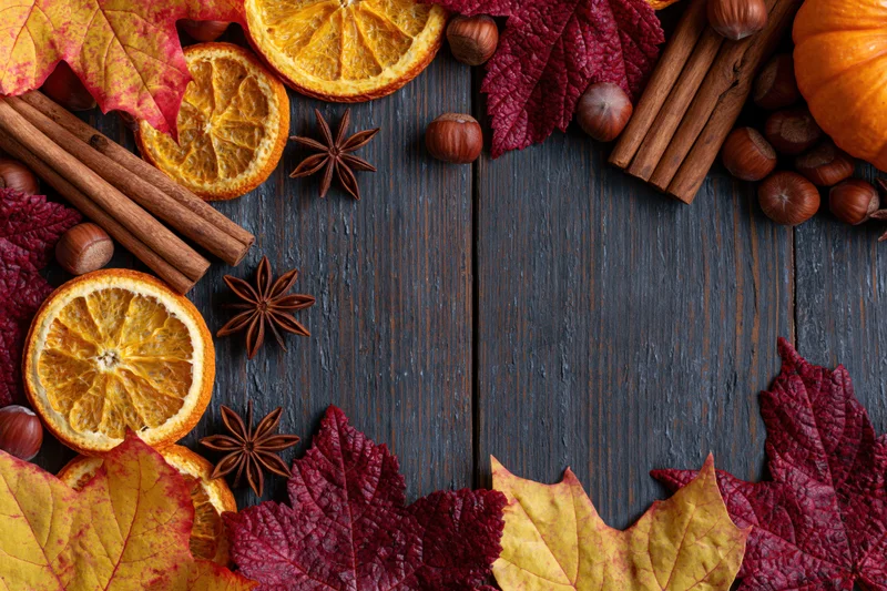 Autumn flat lay arrangement of dried orange slices, cinnamon sticks, star anise, hazelnuts, and colorful maple leaves on dark wooden table with central copyspace