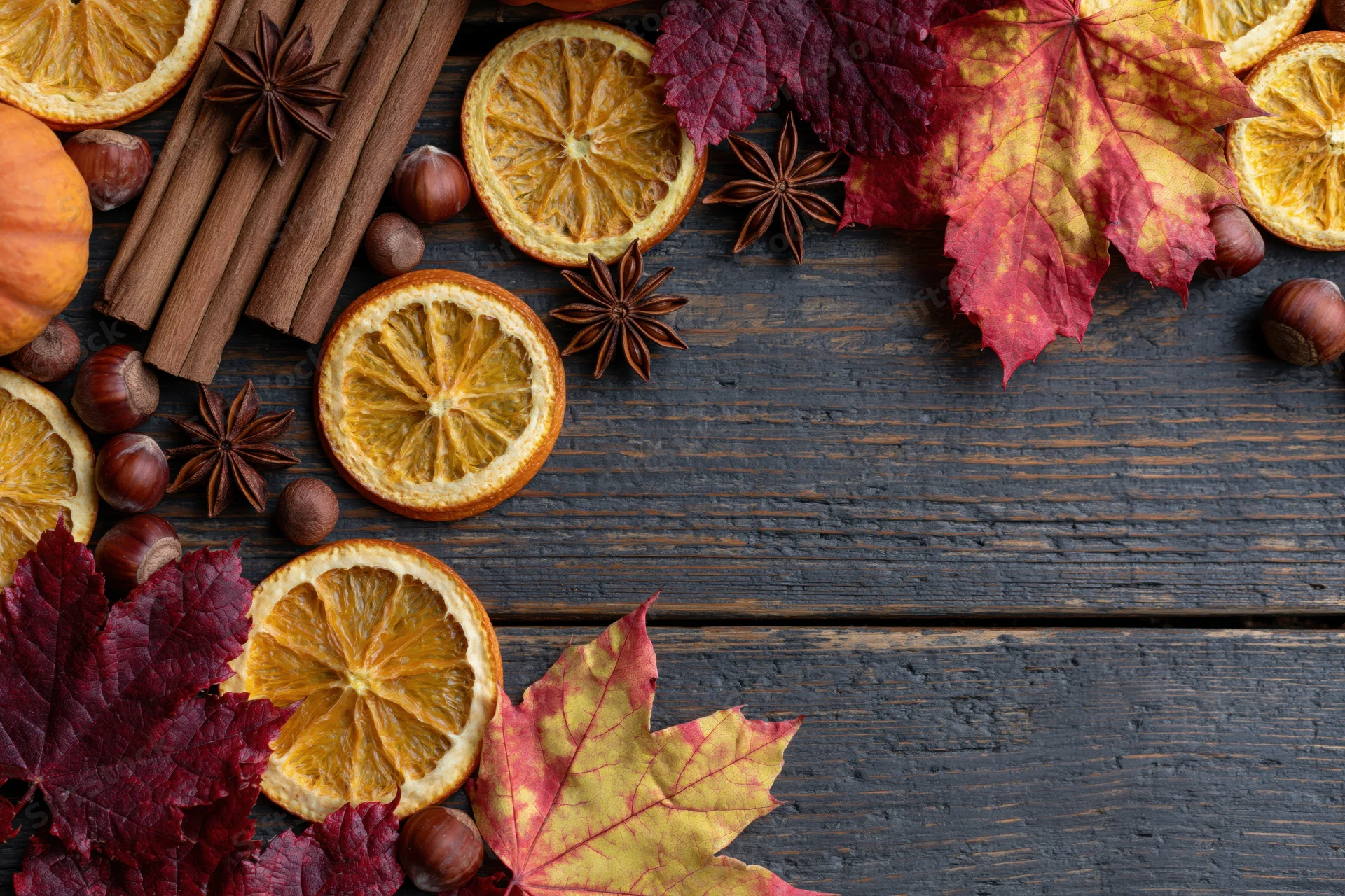Autumn flat lay with dried orange slices, cinnamon sticks, maple leaves, hazelnuts and star anise arranged on dark wooden surface