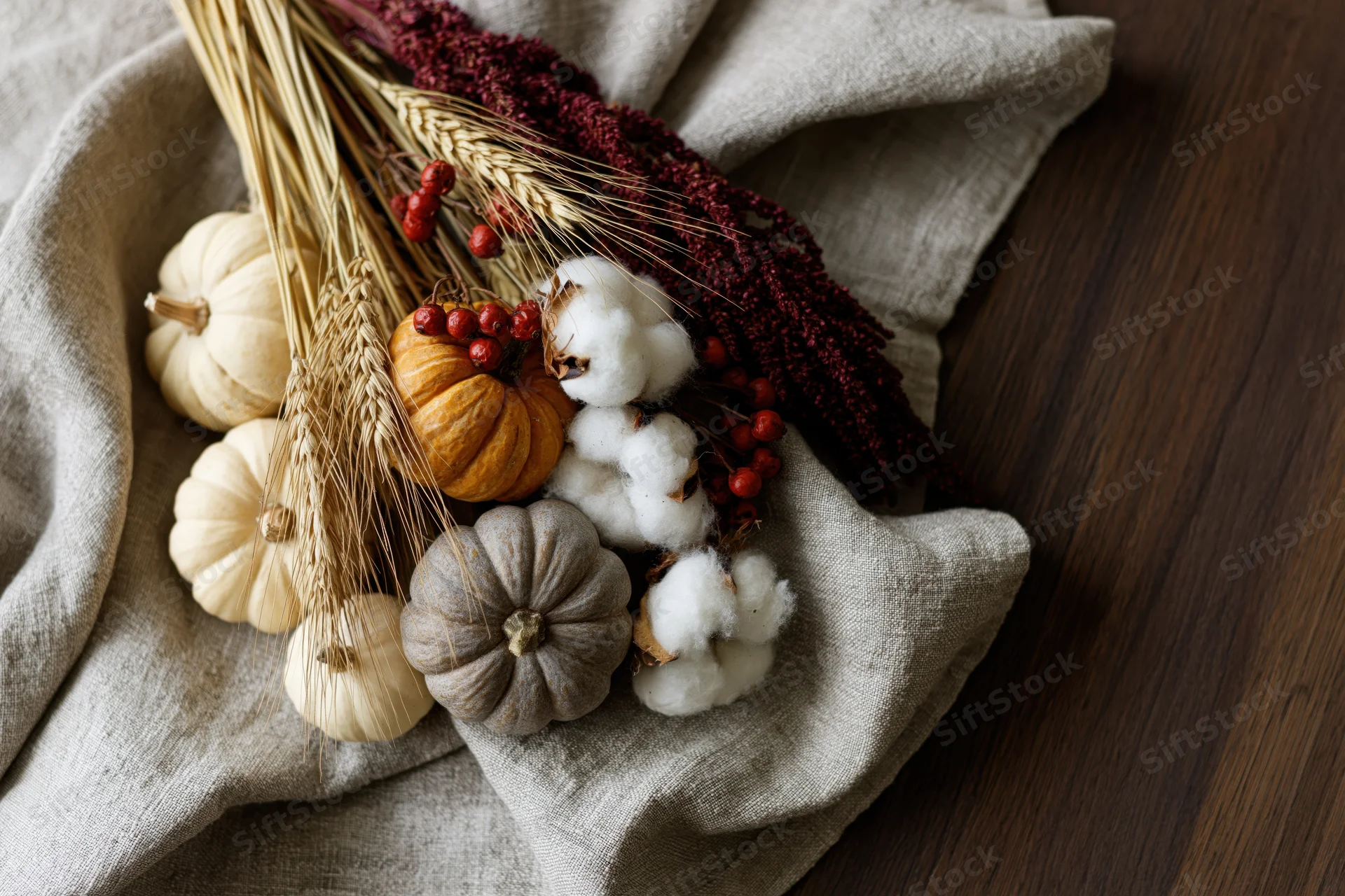 Autumn flat lay with small pumpkins, cotton, wheat, and berries on linen cloth over wooden surface