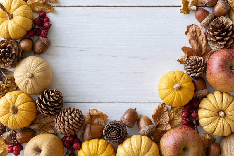 Autumn flat lay arrangement of small yellow and tan pumpkins, pinecones, acorns, apples, and dried leaves on white wooden table with copyspace