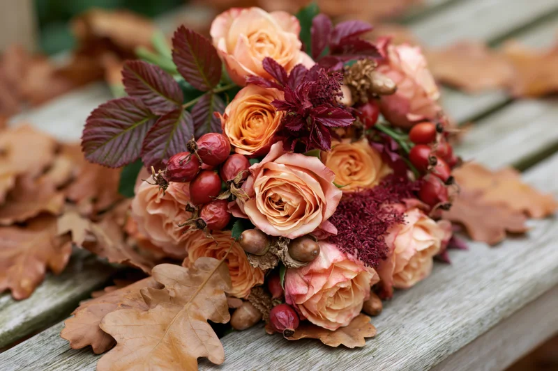 Autumn floral arrangement with roses, rosehips, and oak leaves on a weathered wooden bench