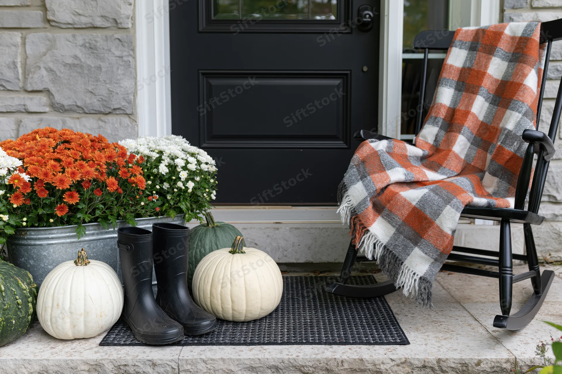 Autumn front porch decorated with white and green pumpkins, orange and white mums, black rainboots, and a plaid blanket on a rocking chair