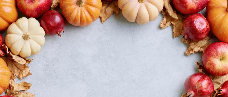 Autumn harvest flatlay arranged as a border frame with pumpkins, apples, pomegranates and dried leaves on a gray surface