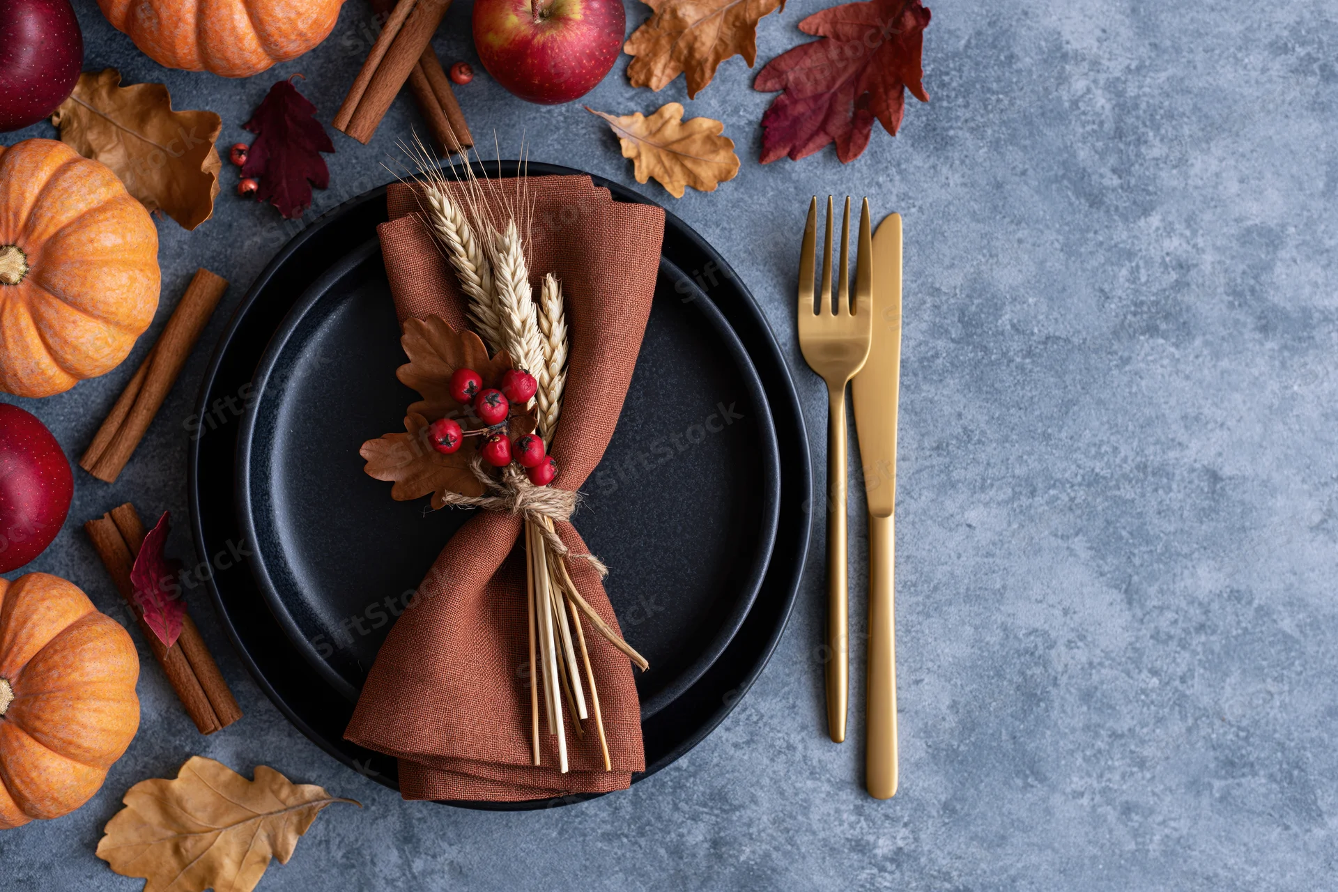 Autumn harvest table setting with pumpkins, apples, cinnamon sticks, and wheat bundle on black plate with gold cutlery, overhead view