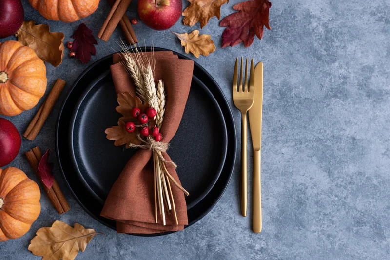 Autumn harvest table setting with pumpkins, apples, cinnamon sticks, and wheat bundle on black plate with gold cutlery, overhead view