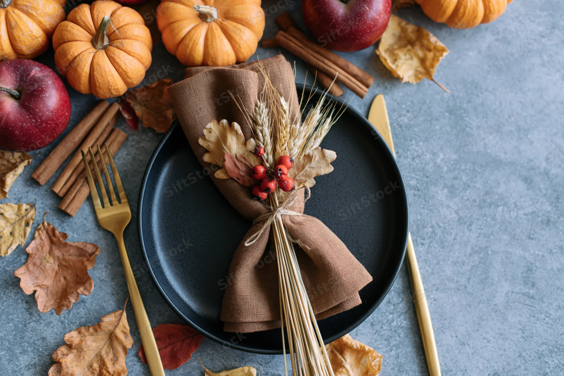 Autumn harvest table setting with pumpkins, apples, cinnamon sticks, and wheat bundle on dark plate with gold cutlery and scattered leaves on gray surface
