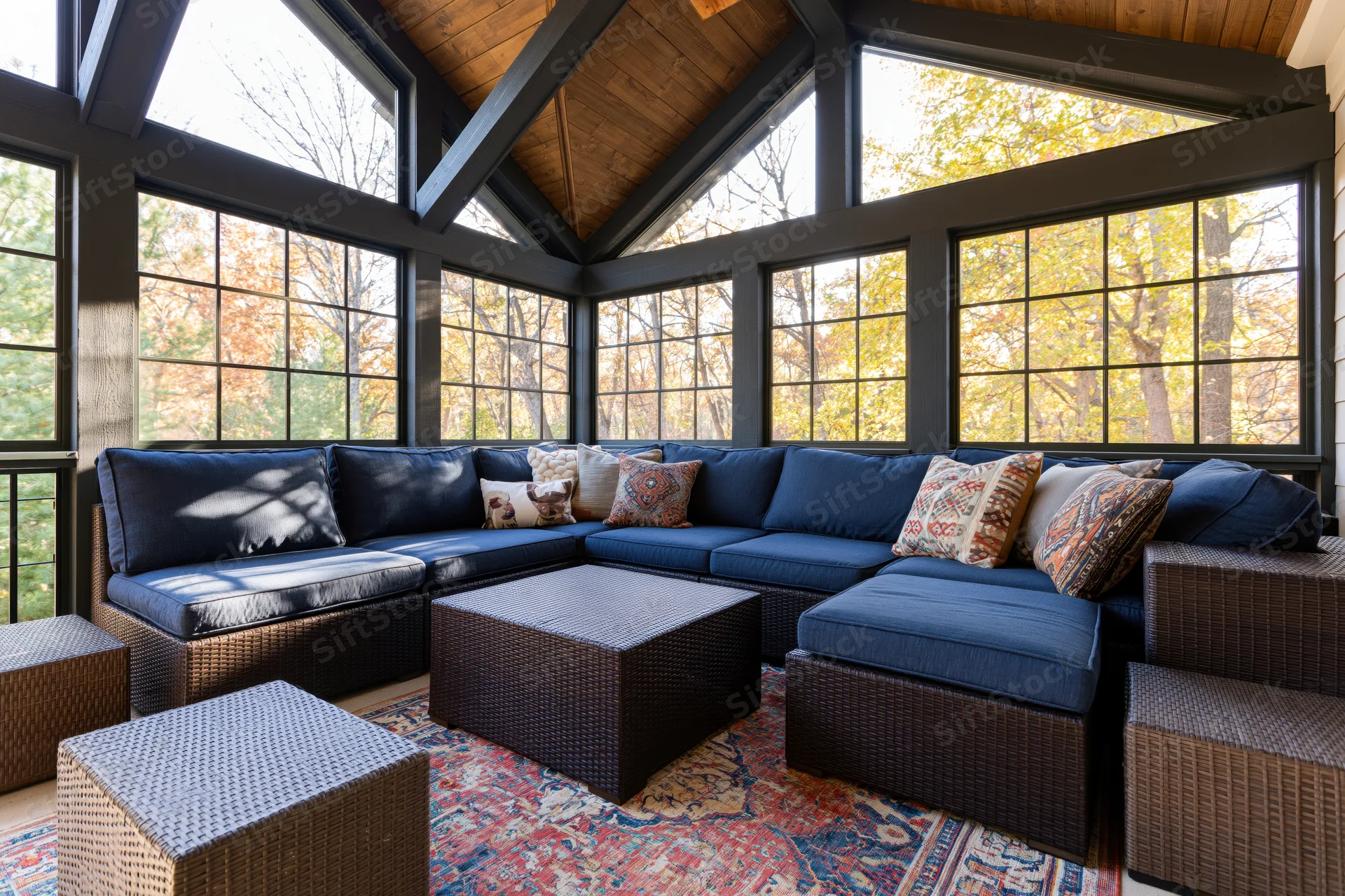 Sectional sofa with blue cushions and patterned pillows arranged in a sunlit screened porch with large windows overlooking autumn trees