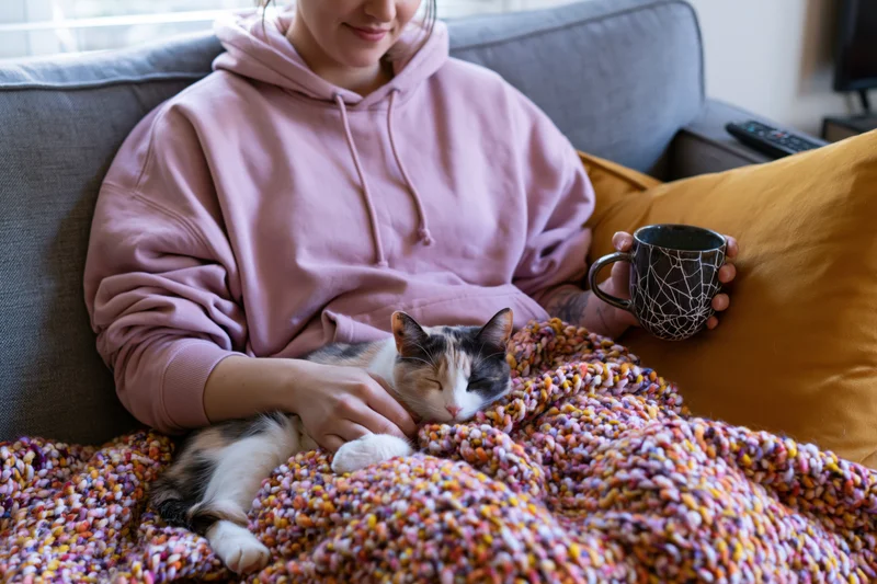 Woman resting on gray sofa with sleeping calico cat under colorful knitted blanket, holding mug