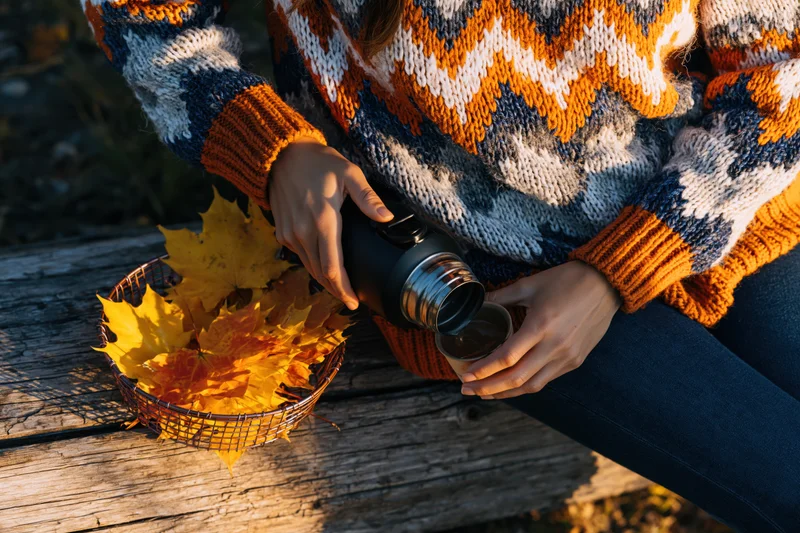 Woman in knitted sweater pouring drink from thermos onto wooden bench beside basket of autumn leaves