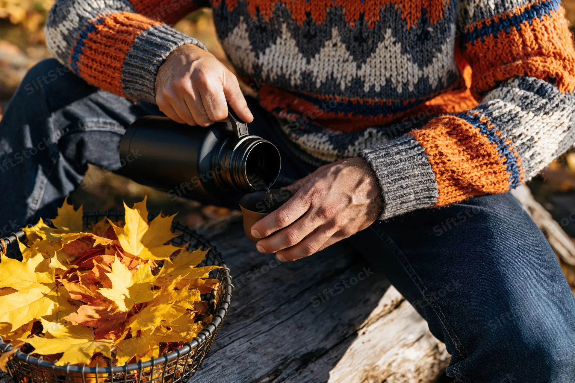 Man pouring coffee from a thermos into a cup while sitting on weathered wood near a basket of autumn leaves