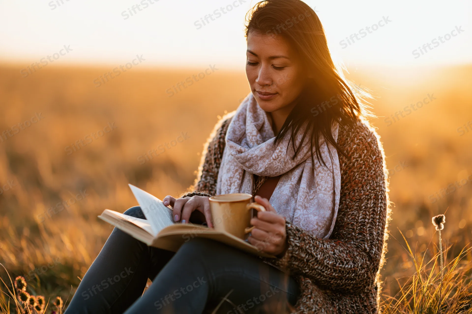 Woman reading a book while holding a mug in a golden field at sunset