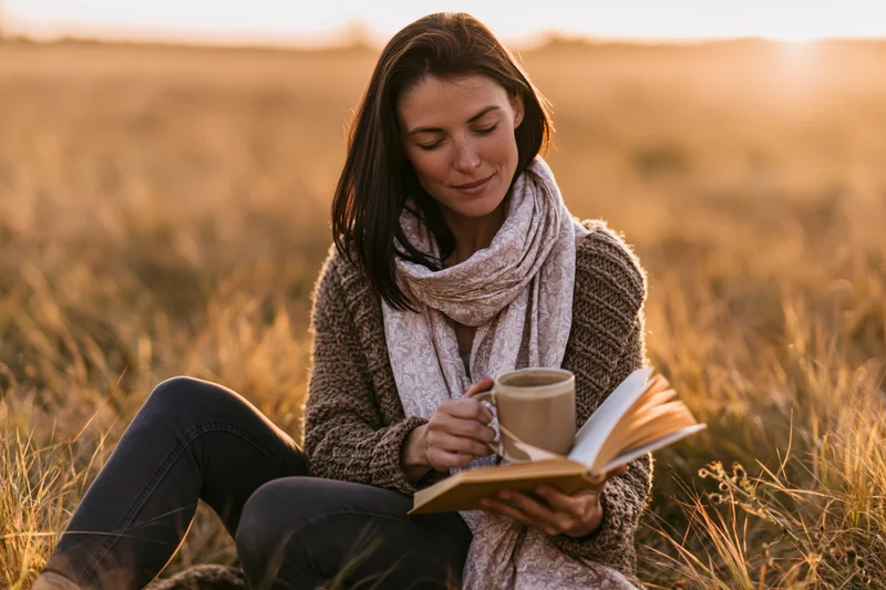 Woman reading a book while holding a mug in a grassy field at sunset