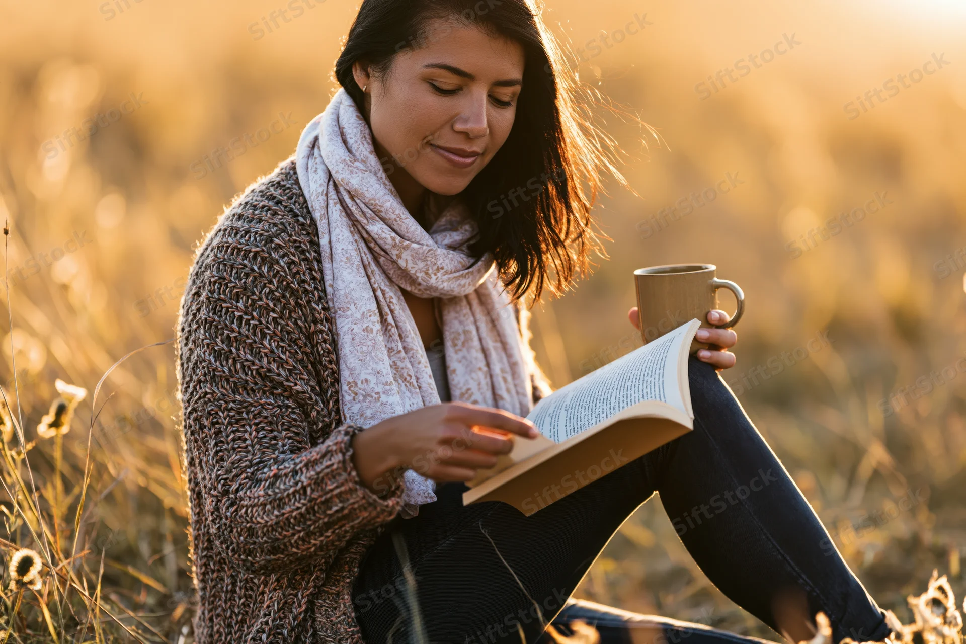 Woman reading a book while holding a mug in a golden field at sunset