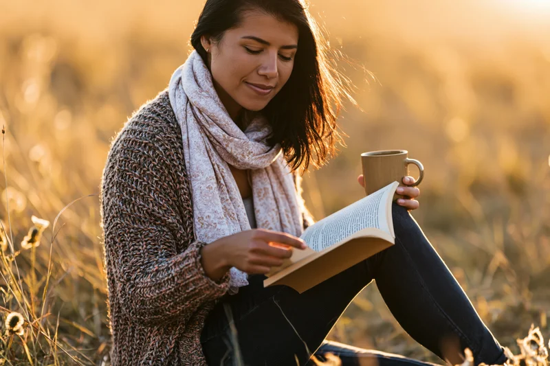 Woman reading a book while holding a mug in a golden field at sunset