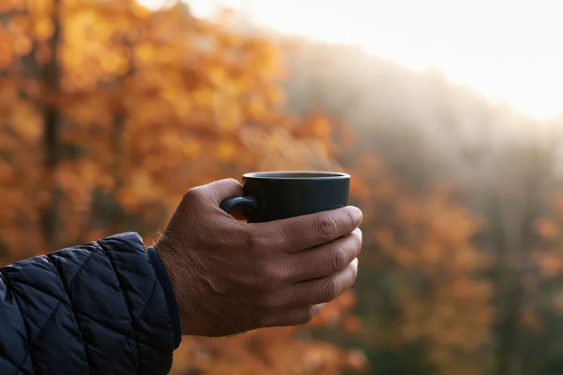 Hand holding a black coffee mug outdoors in an autumn forest at sunrise
