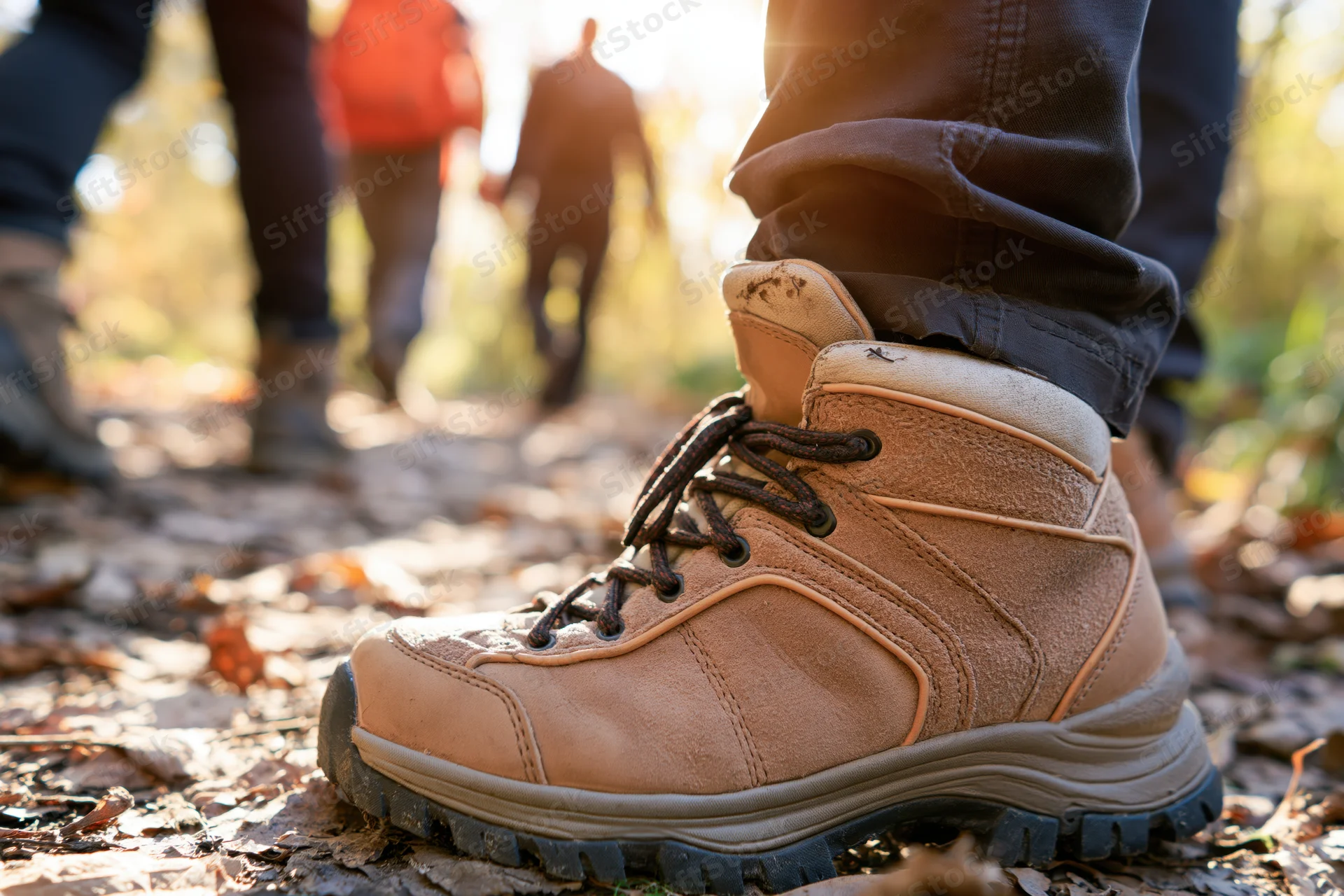 Closeup of a hiking boot on a leaf-covered trail with people walking ahead in a forest
