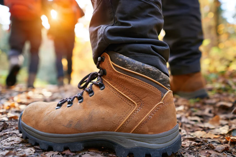 Close-up of hiking boots on a leaf-covered forest trail with sunlight filtering through trees