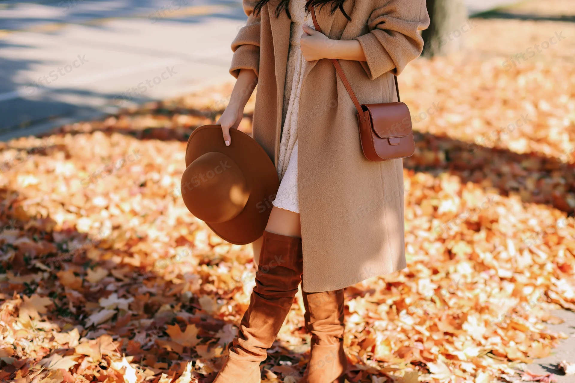 Woman in beige coat and brown boots holding hat while walking on leaf-covered sidewalk