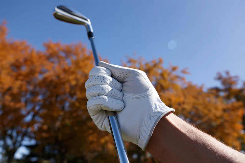 Close-up of a gloved hand gripping a golf club against autumn trees and blue sky