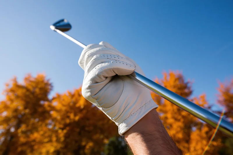 Close-up of a male hand in a white golf glove gripping a golf club against autumn trees and blue sky