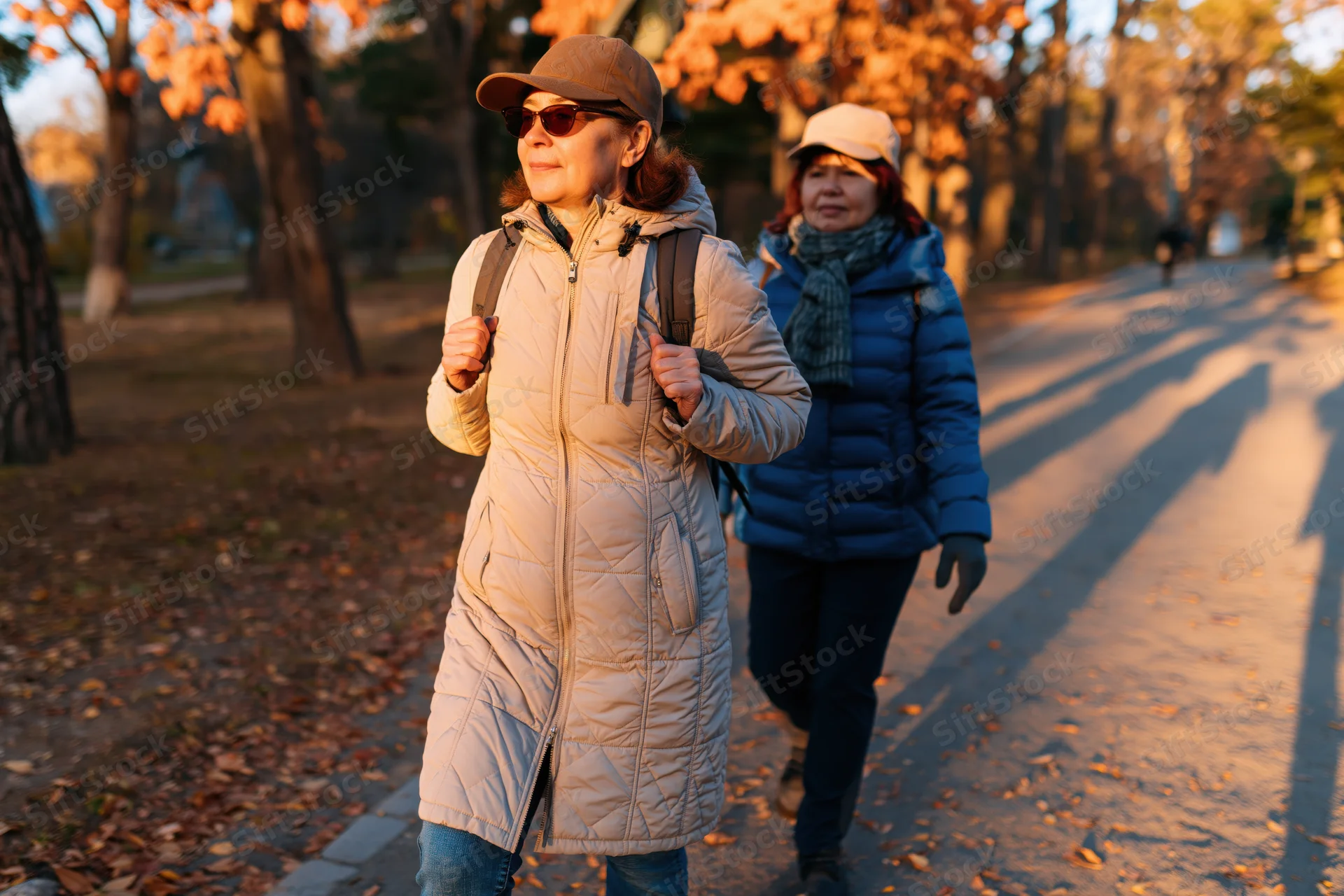 Two women walking along a tree-lined path in a park during autumn golden hour
