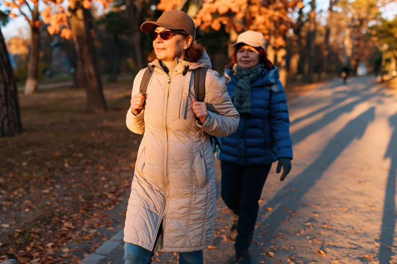 Two women walking along a tree-lined path in a park during autumn golden hour