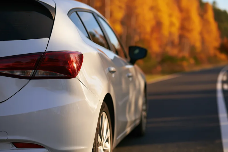 White hatchback driving on a road lined with golden autumn trees