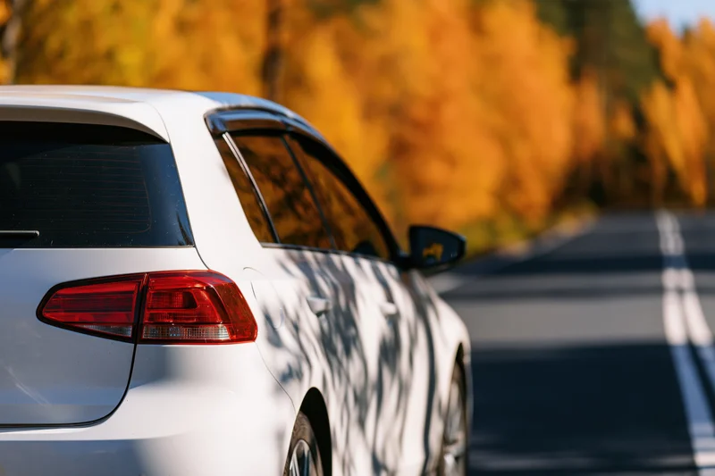 White hatchback driving on a road lined with autumn trees, rear-left view