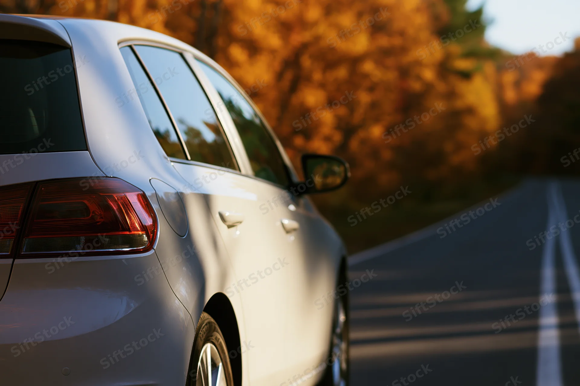 White hatchback car viewed from rear-left side driving on a road lined with autumn trees