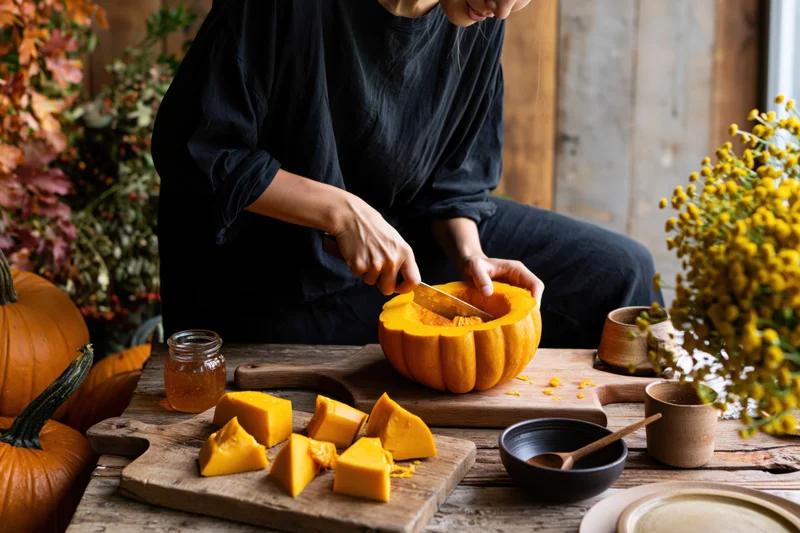 Woman cutting pumpkin on wooden table surrounded by autumn decor and harvest vegetables