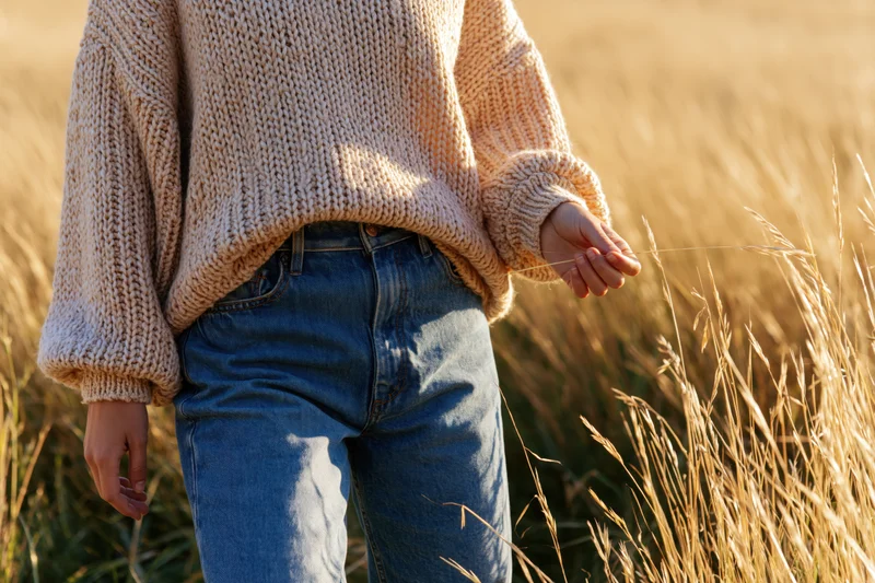 Woman in chunky beige knit sweater and jeans standing in golden wheat field during autumn sunlight