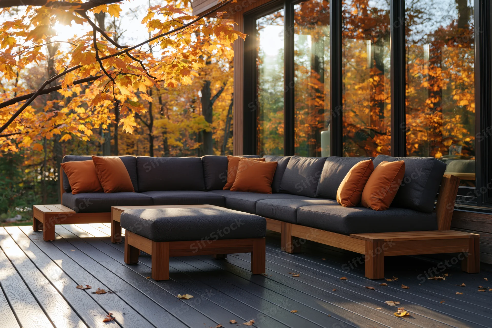 Modern sectional sofa with cushions on a wooden deck beside a glass-walled house surrounded by autumn trees