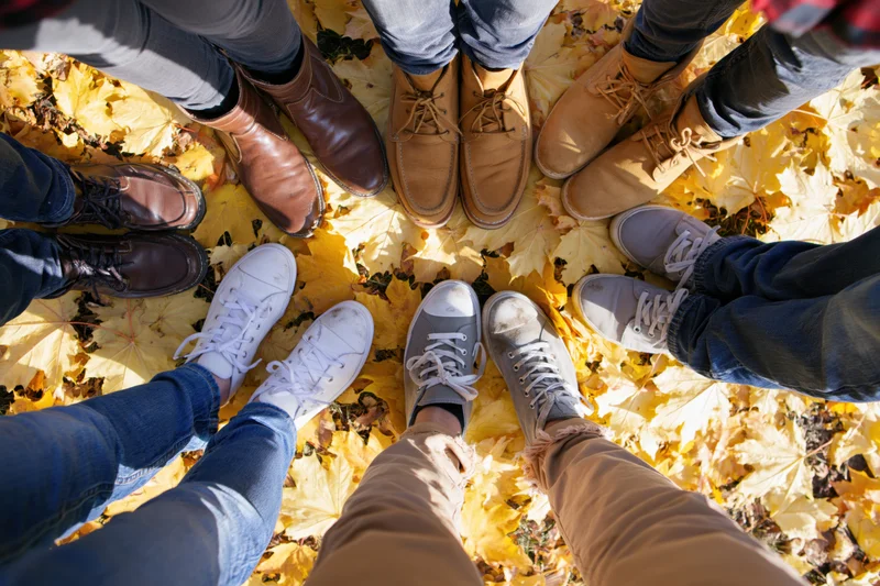 Overhead view of six people standing in a circle on autumn leaves, showing their feet and shoes in casual footwear and jeans
