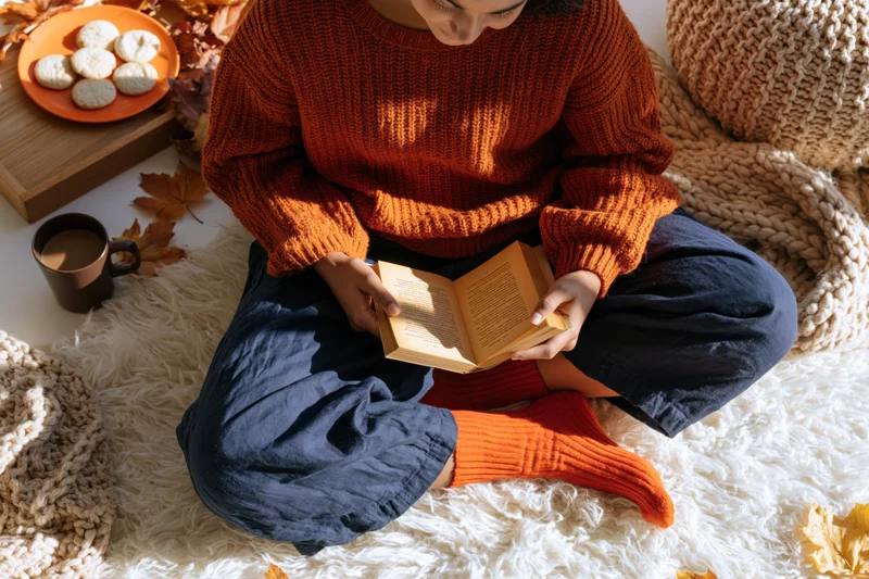 Woman reading a book while sitting on a white rug surrounded by autumn leaves and cozy items