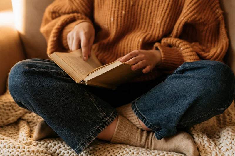 Woman reading a book while sitting cross-legged on a couch covered with a knitted blanket